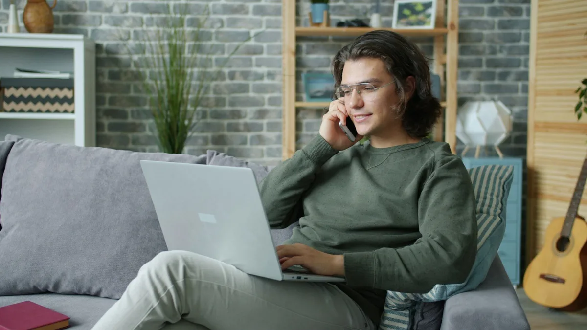 Mosolygó fiatal férfi telefonál, közben a laptopján dolgozik otthon