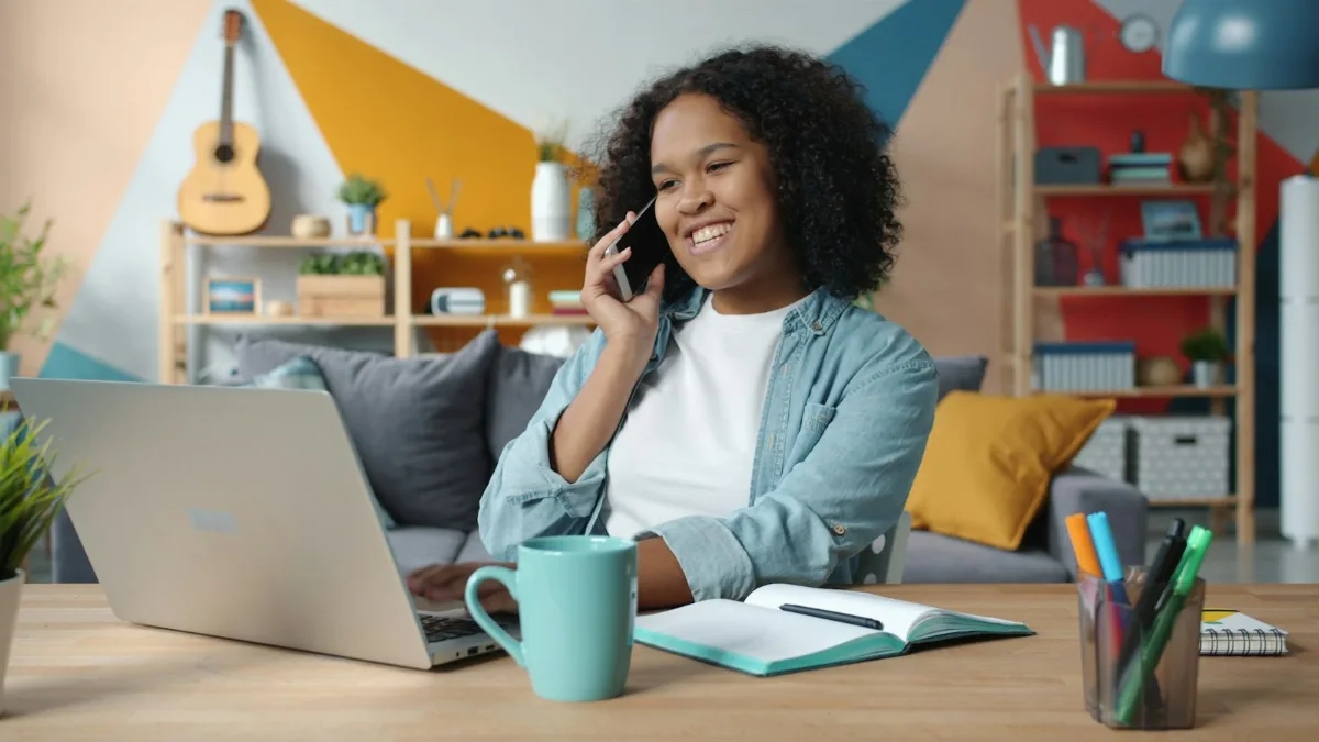 Mosolygó nő telefonál, közben az íróasztalánál a laptopján dolgozik